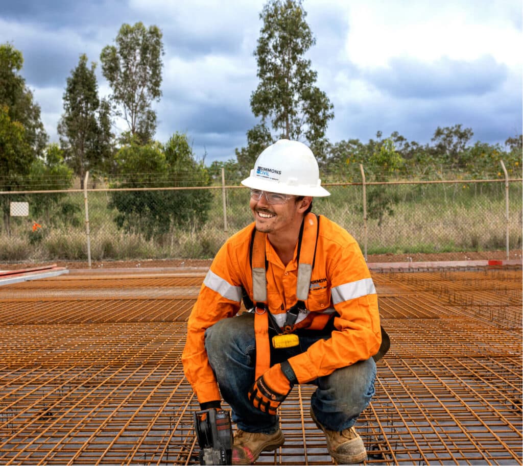 Simmons worker in orange work shirt and hard hat crouching and smiling